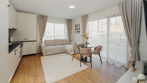 A modern living room with a sofa, a dining table with chairs, a kitchen area, and large windows with curtains.
