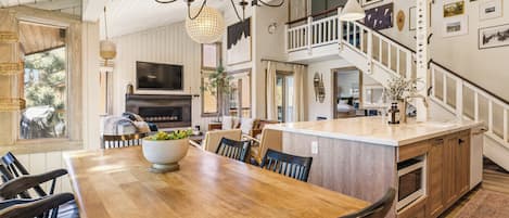 A spacious dining area with a wooden table and chairs, a large bowl of fruit, and a television above the fireplace.