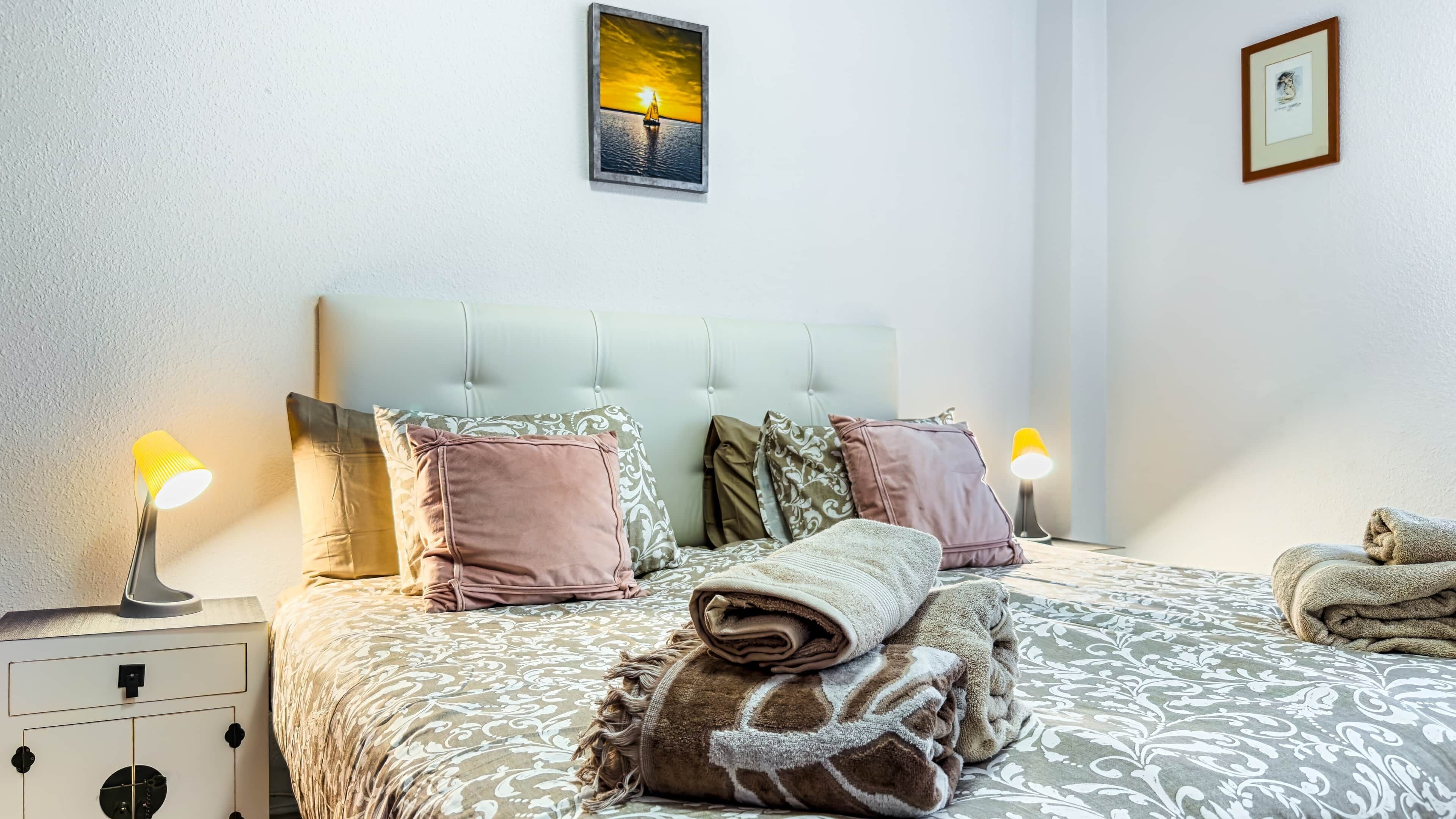 Een net opgemaakt bed met een beddekleed met dessin, kussens en een opgerolde deken. Twee nachtlampen, een nachtkastje met lamp en een ingelijste foto aan de muur.