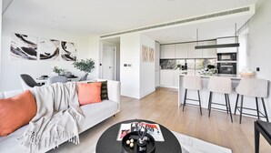 A modern living room with a sofa, coffee table, and artwork. A kitchen with bar stools and appliances is visible in the background. - Classy Modern Flat | Luxury & Comfy | Near Westfield & Tube (Greater London)