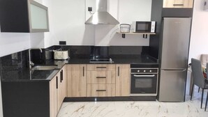A modern kitchen with black countertops, wooden cabinets, a stainless steel range hood, a microwave, a toaster, and a refrigerator. - PORTOCEL I (Marín)
