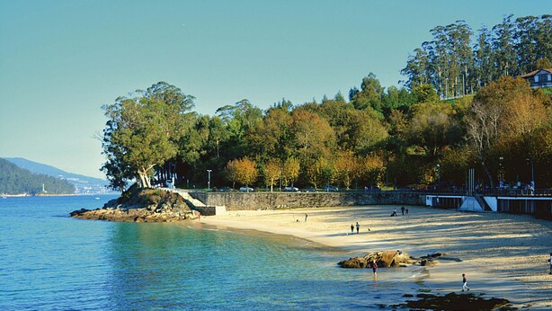 A beach with clear water, a rocky shoreline, and a forested hill in the background. - PORTOCEL I (Marín)
