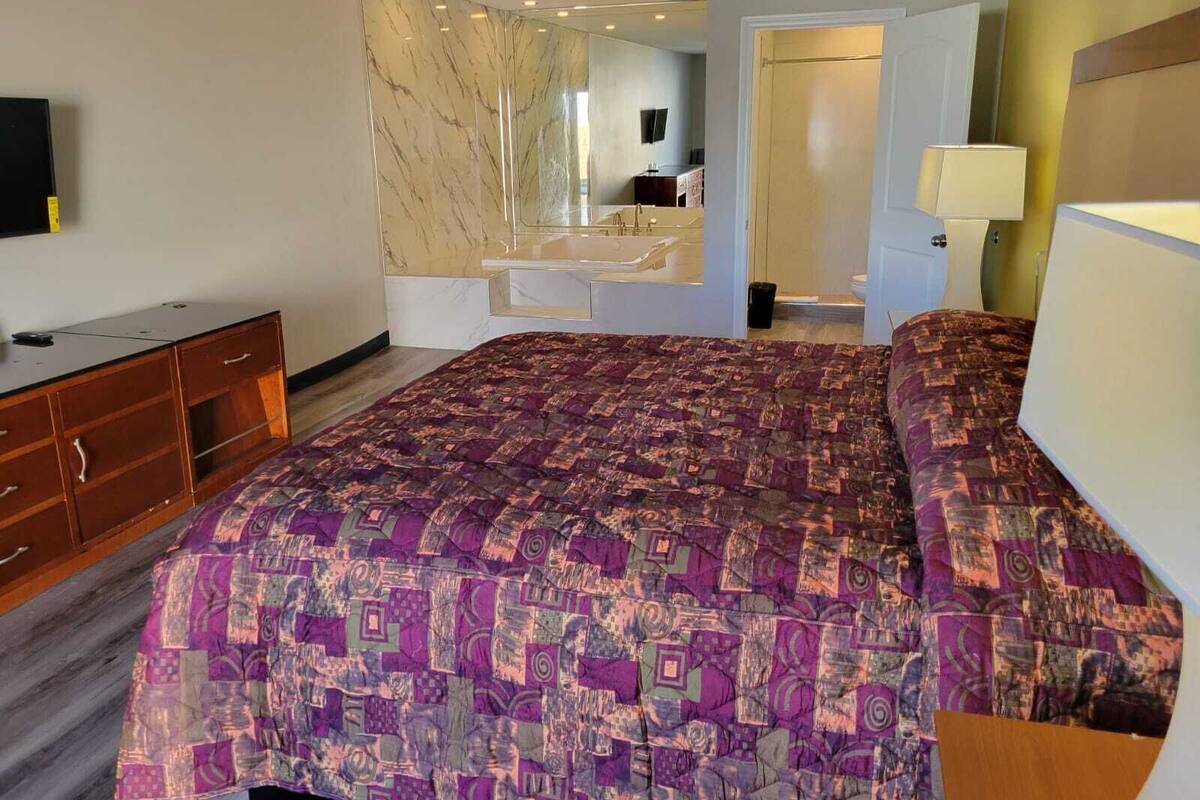 A bedroom with a large bed, a wooden dresser, a mirror, and a bathroom visible through an open door.