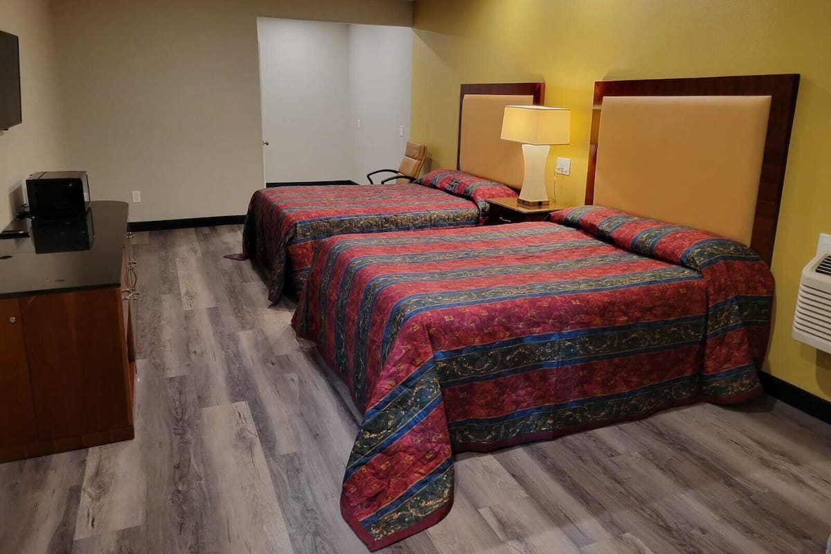 A hotel room with two beds, a desk, a chair, a lamp, and a wall-mounted air conditioner.