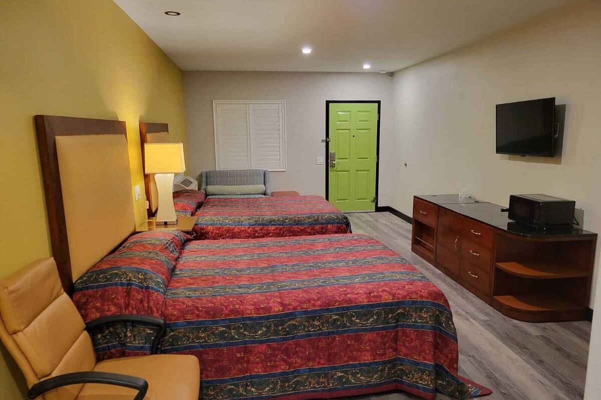 A hotel room with two beds, a chair, a desk with a TV, and a green door.