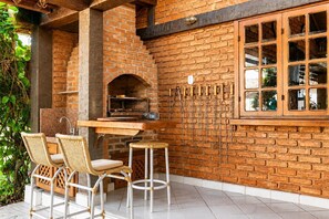 An outdoor kitchen area with a brick oven, bar stools, and hanging kitchen tools. - Bz46 Casa 150 metros da praia 5 suites (Buzios)