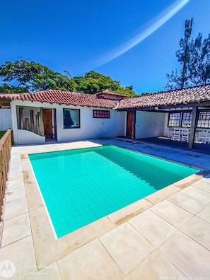A swimming pool with tiled flooring, a covered patio area, and a building with a tiled roof. - Bz46 Casa 150 metros da praia 5 suites (Buzios)