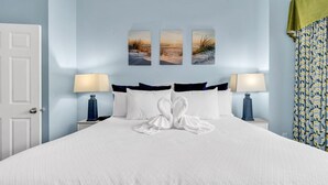 Een net opgemaakt bed met een witte deken en kussens, twee bedlampen en drie aan de muur bevestigde schilderijen met strandtaferelen.