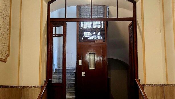 Una amplia escalera con barandilla de madera que conduce a una puerta con panel de vidrio y reja metálica.