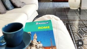 Un salón con un sofá blanco, una mesa de centro con un libro titulado 'ROME', una taza azul y una alfombra estampada.