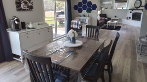 A dining area with a table covered in plastic, surrounded by chairs. A white cabinet with drawers is against the wall, and a window with curtains is visible. - Beautiful, cozy, spacious suite located central PEI. 3 bedrooms 4 beds.  (Lot 67)