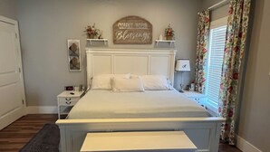A neatly arranged bedroom with a bed, bedside tables, a nightstand, a bedside lamp, a decorative sign above the bed, and a window with curtains. - Family-friendly close to SDC, Branson, Eureka Springs, Table Rock, Roaring River (Cassville)