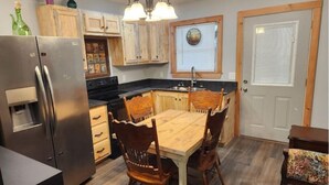 A compact kitchen with wooden cabinets, a stainless steel refrigerator, a dining table with four chairs, and a window with blinds. - Cedar Grove Rental A-frame (Clarksville)