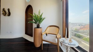 Habitación de esquina con piso de madera oscura, una silla de mimbre, un helecho en maceta, una pequeña mesa redonda con una taza y platillo, y vistas a edificios a través de un gran ventanal.