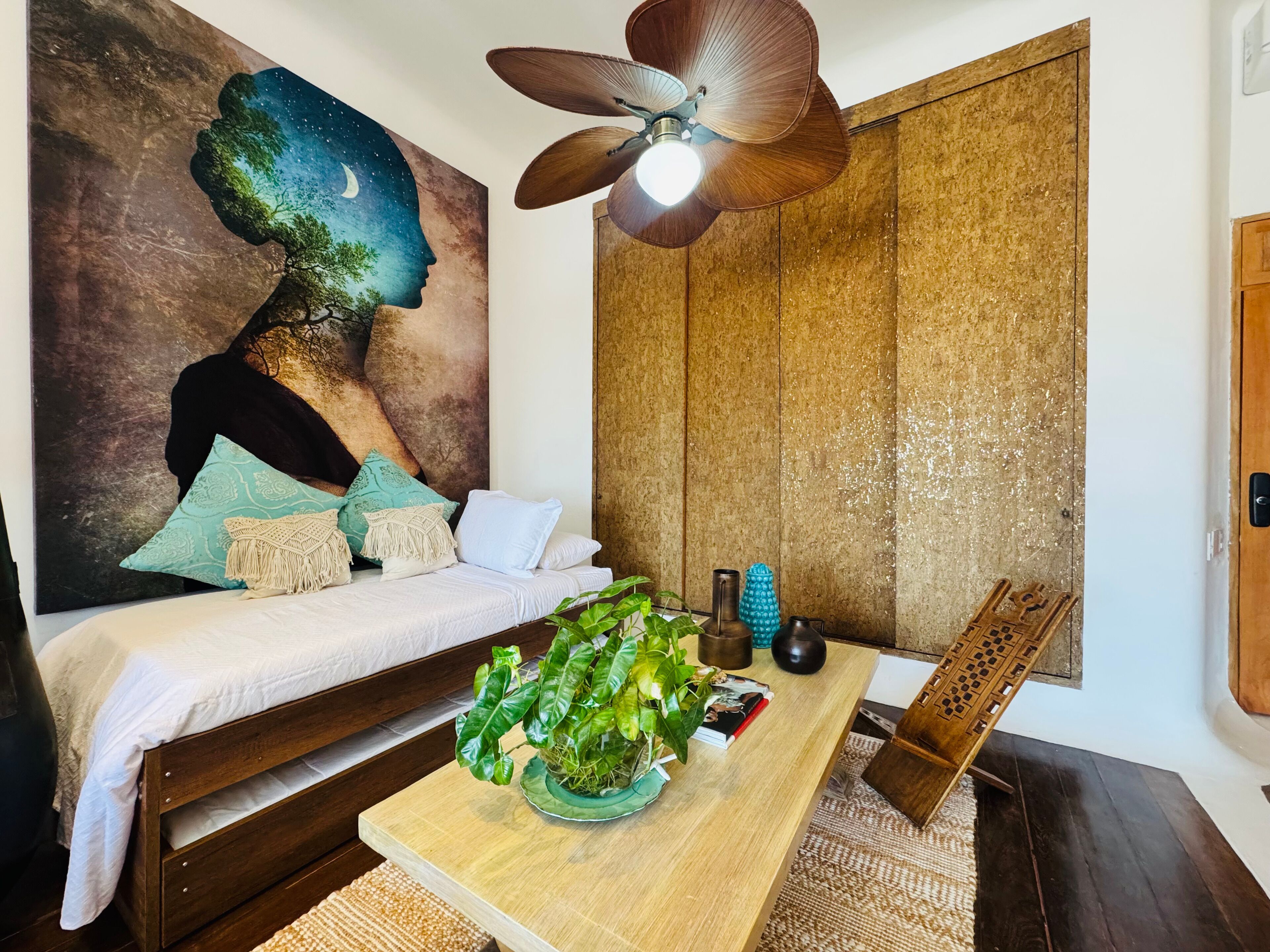 Une chambre à coucher comprenant un lit, une table de chevet en bois, une table avec des plantes, un ventilateur de plafond fixé au mur et un grand tableau abstrait.