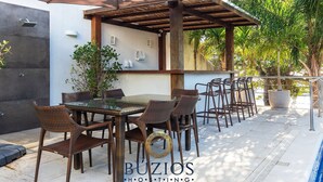 An outdoor dining area with wicker chairs and a glass table, a pergola, and a poolside setting.