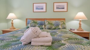 Ein ordentlich bezogenes Bett mit Bettwäsche mit tropischem Blattmuster, zwei Nachttischlampen und zwei gerahmte Bilder an der Wand.