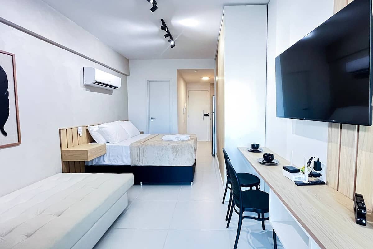 Quarto de hotel moderno com cama, sofá, televisão na parede e uma pequena mesa com cadeiras.