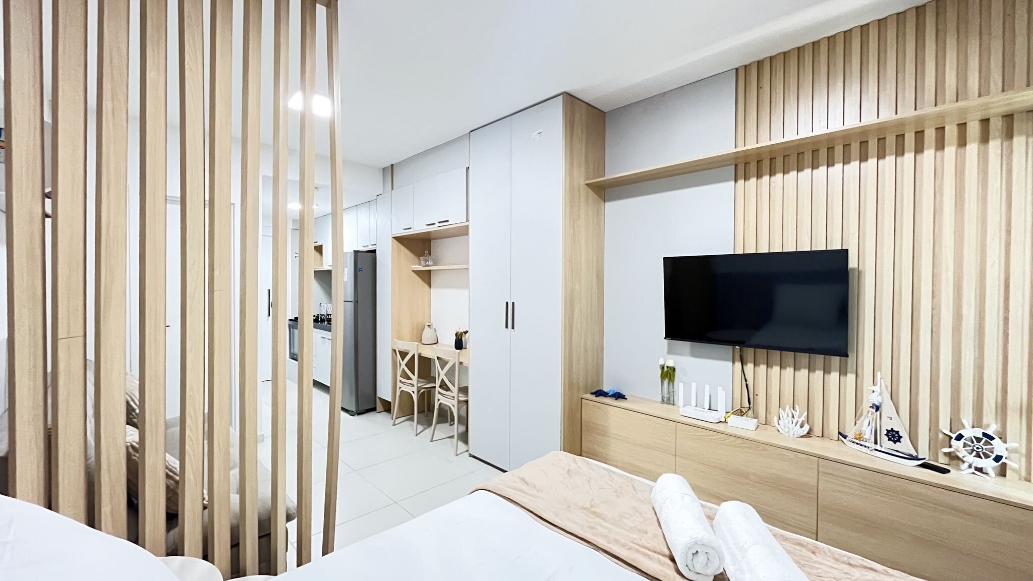 A modern hotel room with a bed, a TV mounted on a wooden wall, a desk with a chair, and a kitchenette in the background.