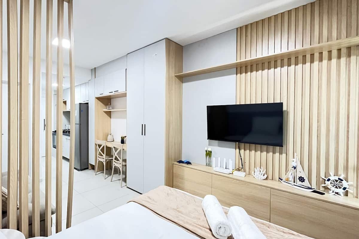Quarto de hotel moderno com uma cama, uma TV fixada numa parede de madeira, uma escrivaninha com cadeira e uma kitchenette ao fundo.