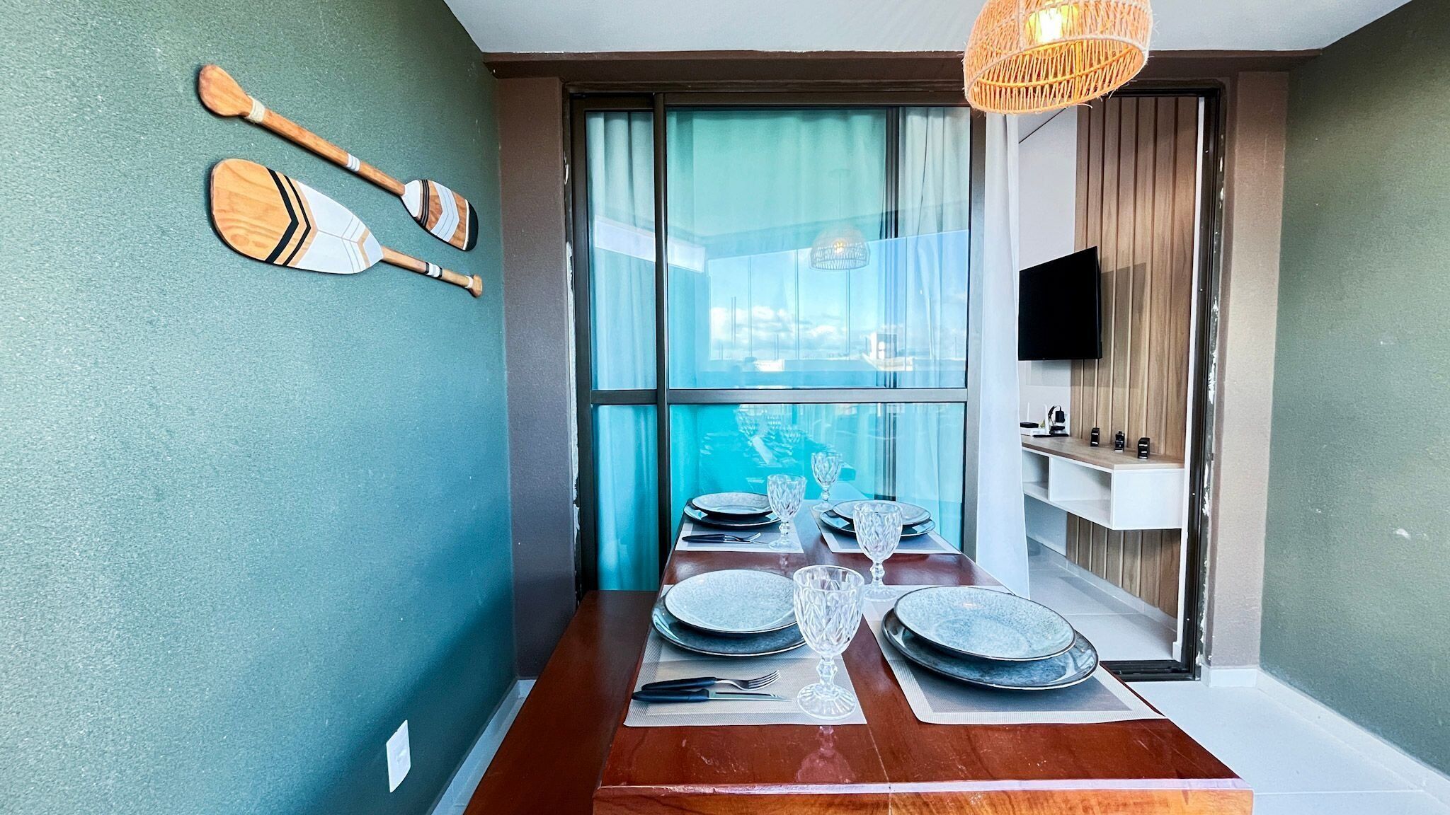 A dining area with a wooden table set for four, a television mounted on the wall, and a view through a large window.