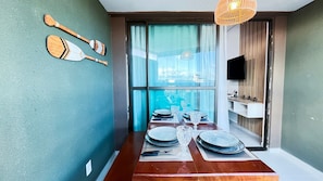 A dining area with a wooden table set for four, a television mounted on the wall, and a view through a large window. - Açores Tamandaré Beach Beira-Mar (Tamandare)