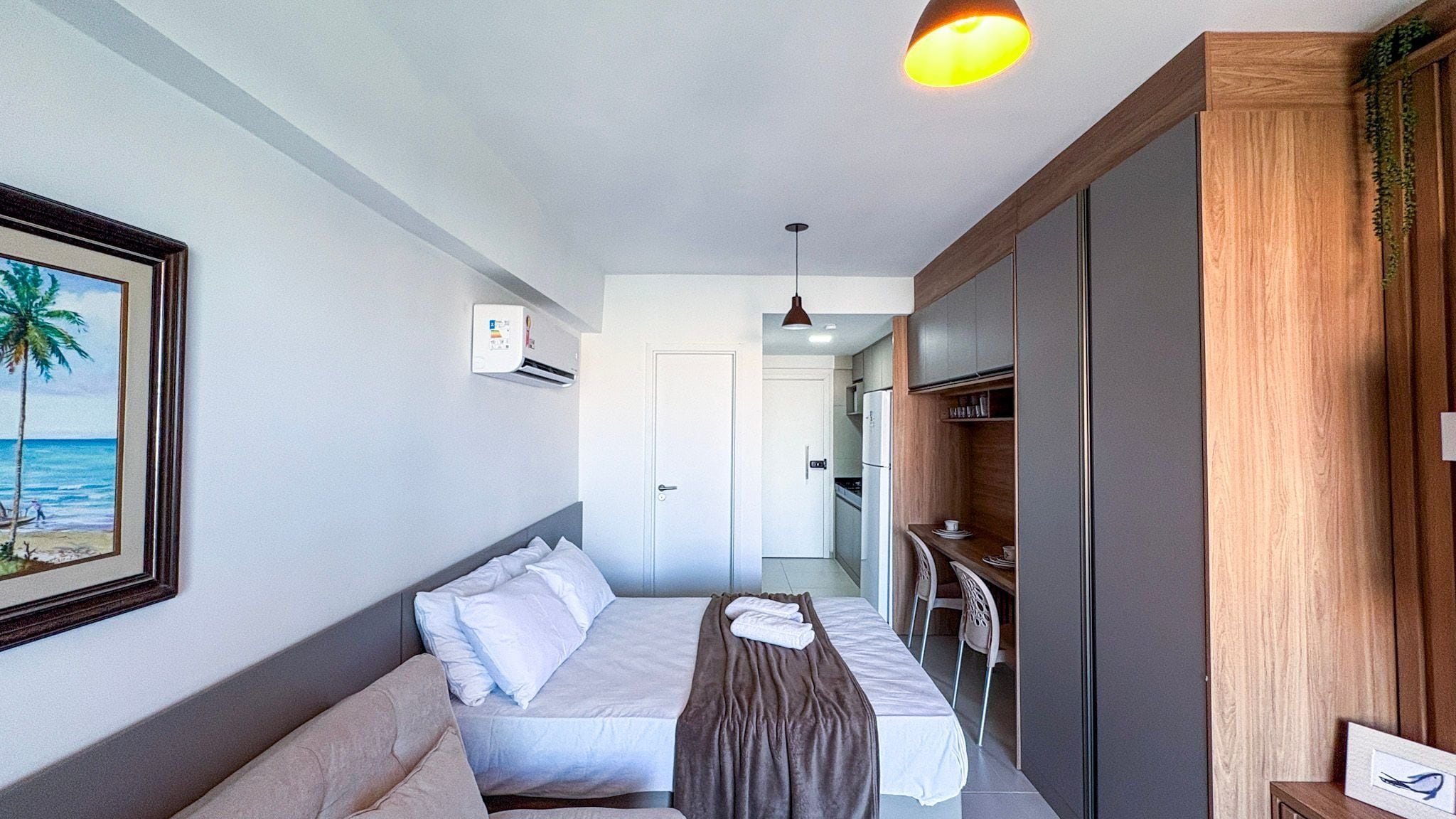 Et hotelværelse med seng, sengebord, et billede i ramme på væggen, en aircondition og udsigt til det tilstødende køkken.