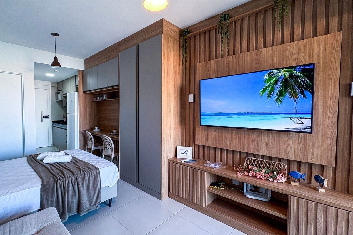 Habitación de hotel con cama, televisor montado en la pared, kitchenette y paneles de madera.