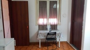 Pequeña habitación con puerta de madera, ventana con cortinas, una mesa, una silla y piso de baldosas.