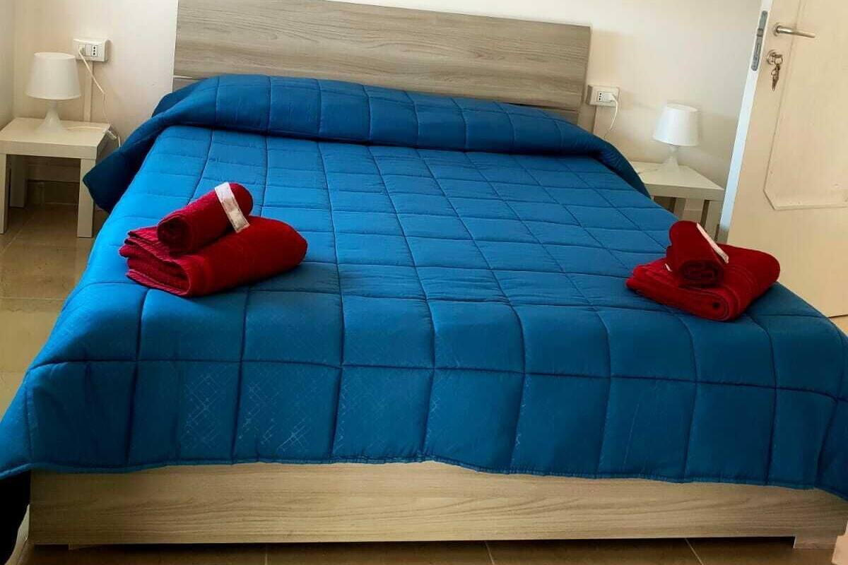 Una cama bien hecha con colcha azul y dos toallas rojas, cabecera de madera y lámparas de noche sobre mesitas blancas.