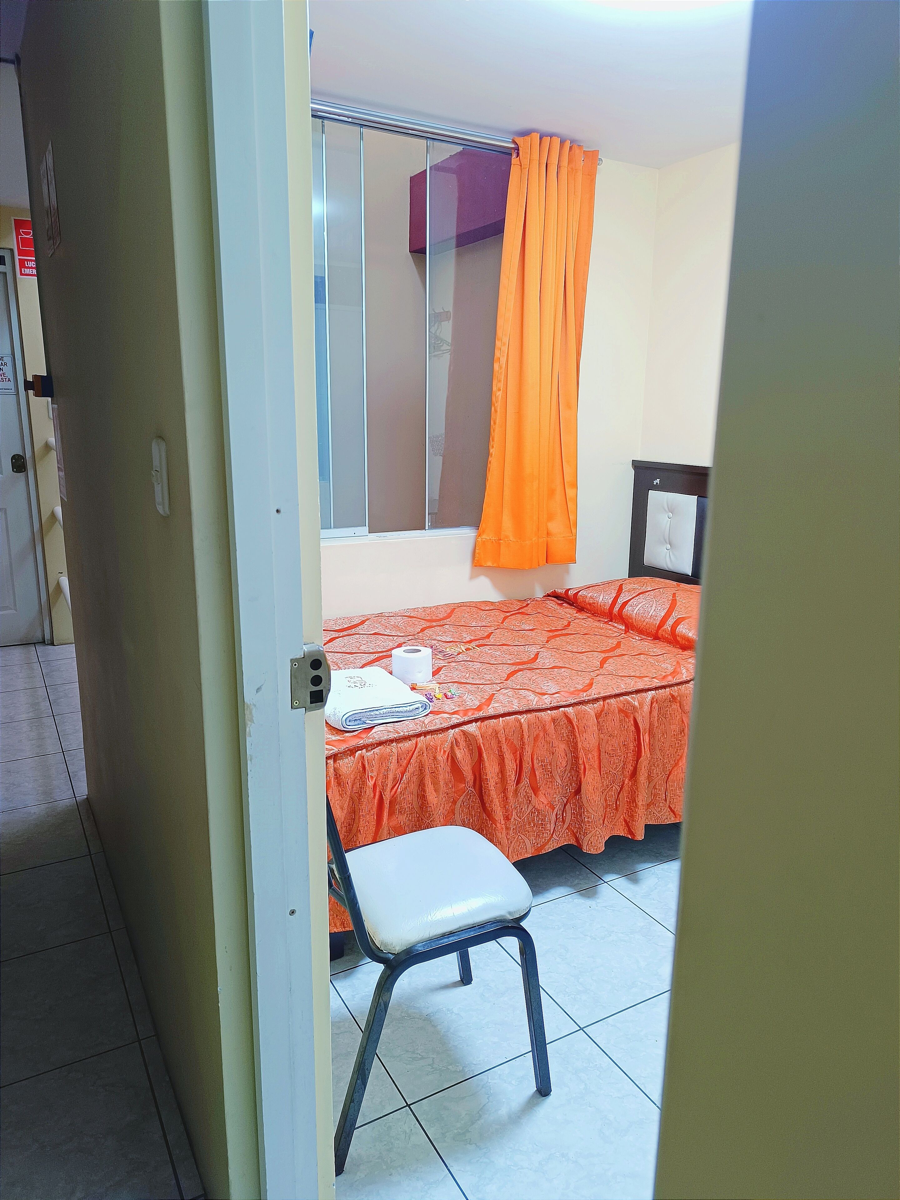 Une chambre d’hôtel avec un lit, une chaise, une fenêtre avec des rideaux orange et un miroir.