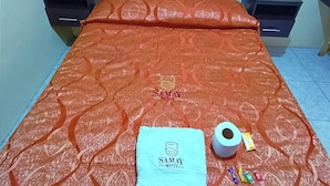 Ein ordentlich bezogenes Bett mit einer orangefarbenen, gemusterten Tagesdecke, einem blauen Handtuch und einer Toilettenpapierrolle.