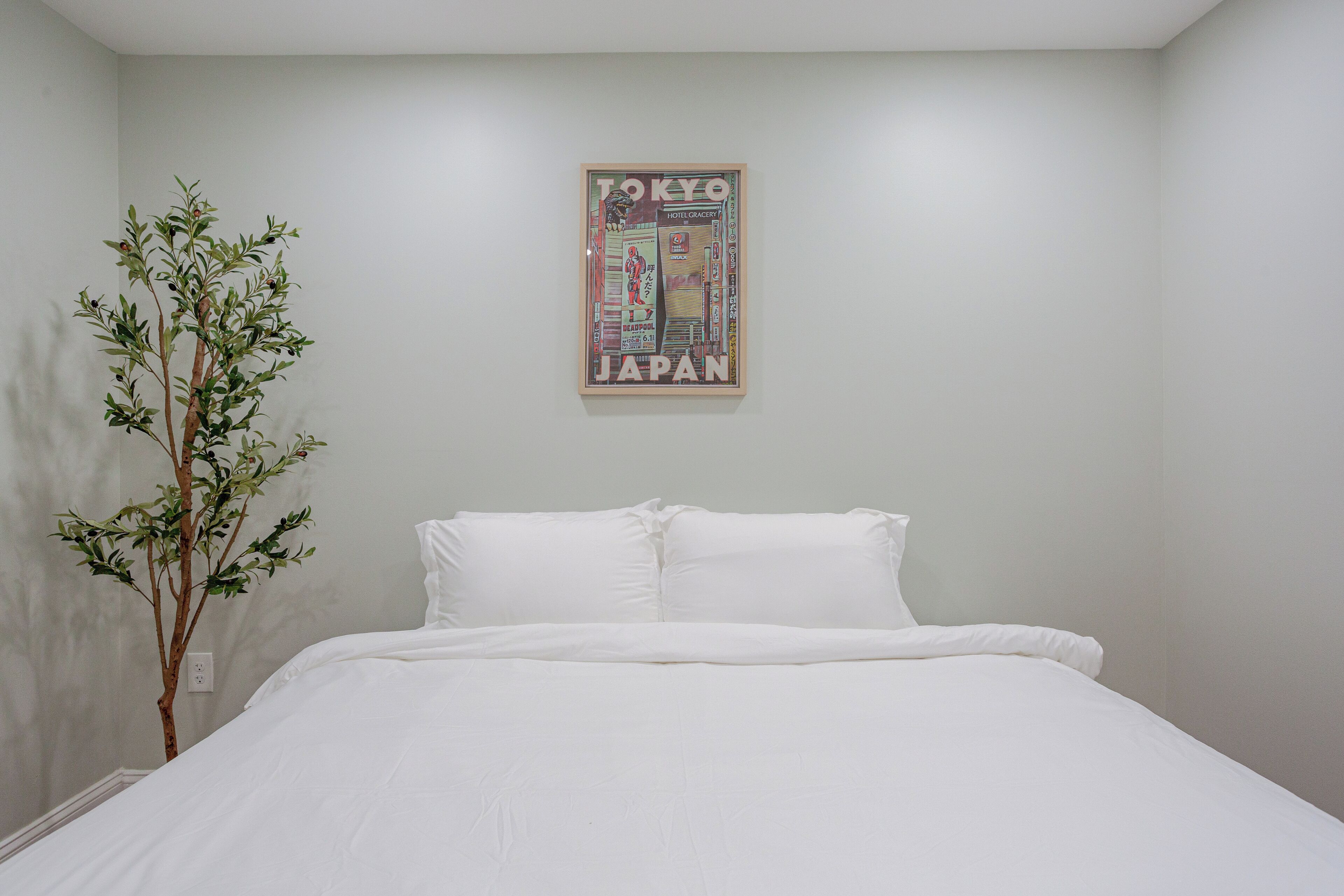 A bedroom with a bed, white bedding, a wall-mounted poster of Tokyo, Japan, and a small tree.