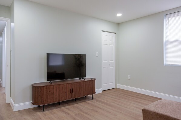 Quarto com televisão de tela plana sobre um móvel de madeira, porta branca e janela com persianas.