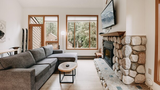 Een moderne woonkamer met een modulaire bank, een stenen open haard en grote ramen met uitzicht op bomen.