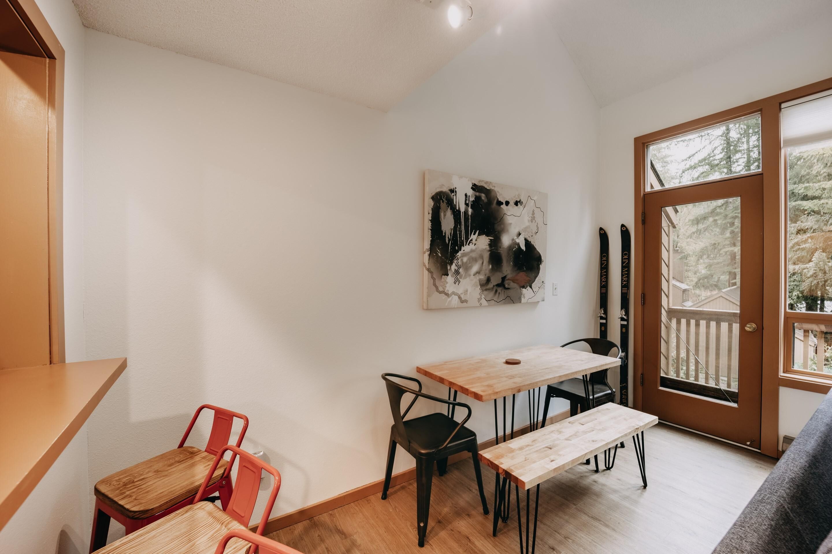 Et lille værelse med et træbord og stole, en bænk, et billede på væggen og en dør til en balkon med udsigt over træer.