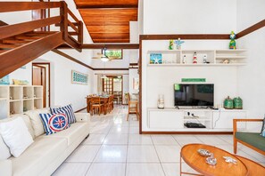 Un vaste espace de vie comprenant un escalier en bois, une table à manger et un téléviseur posé sur une console blanche.