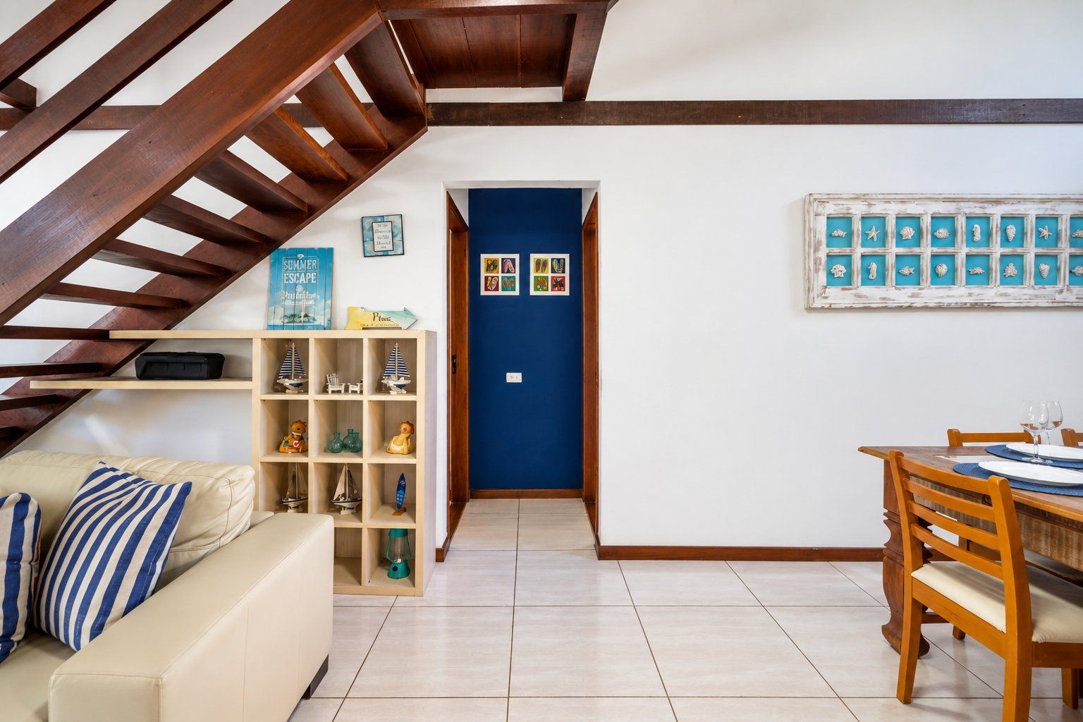 A room with a wooden staircase, a blue door, a dining table with chairs, and a shelf with decorative items.