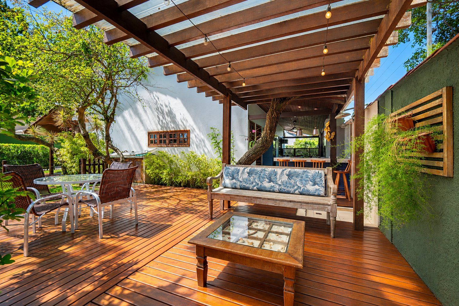 An outdoor patio with wooden flooring, a glass-top table, wicker chairs, a wooden bench with a blue cushion, and a hanging plant.