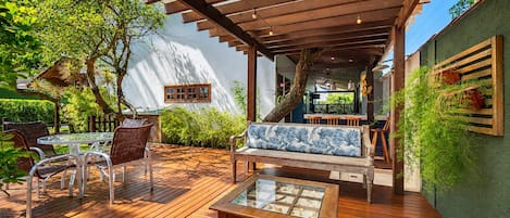 An outdoor patio with wooden flooring, a glass-top table, wicker chairs, a wooden bench with a blue cushion, and a hanging plant.