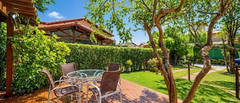 An outdoor patio with a glass table and wicker chairs, surrounded by greenery and a wooden deck.