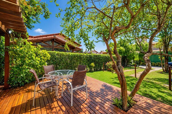 Un patio al aire libre con una mesa de vidrio y sillas de mimbre, rodeado de follaje y una terraza de madera.
