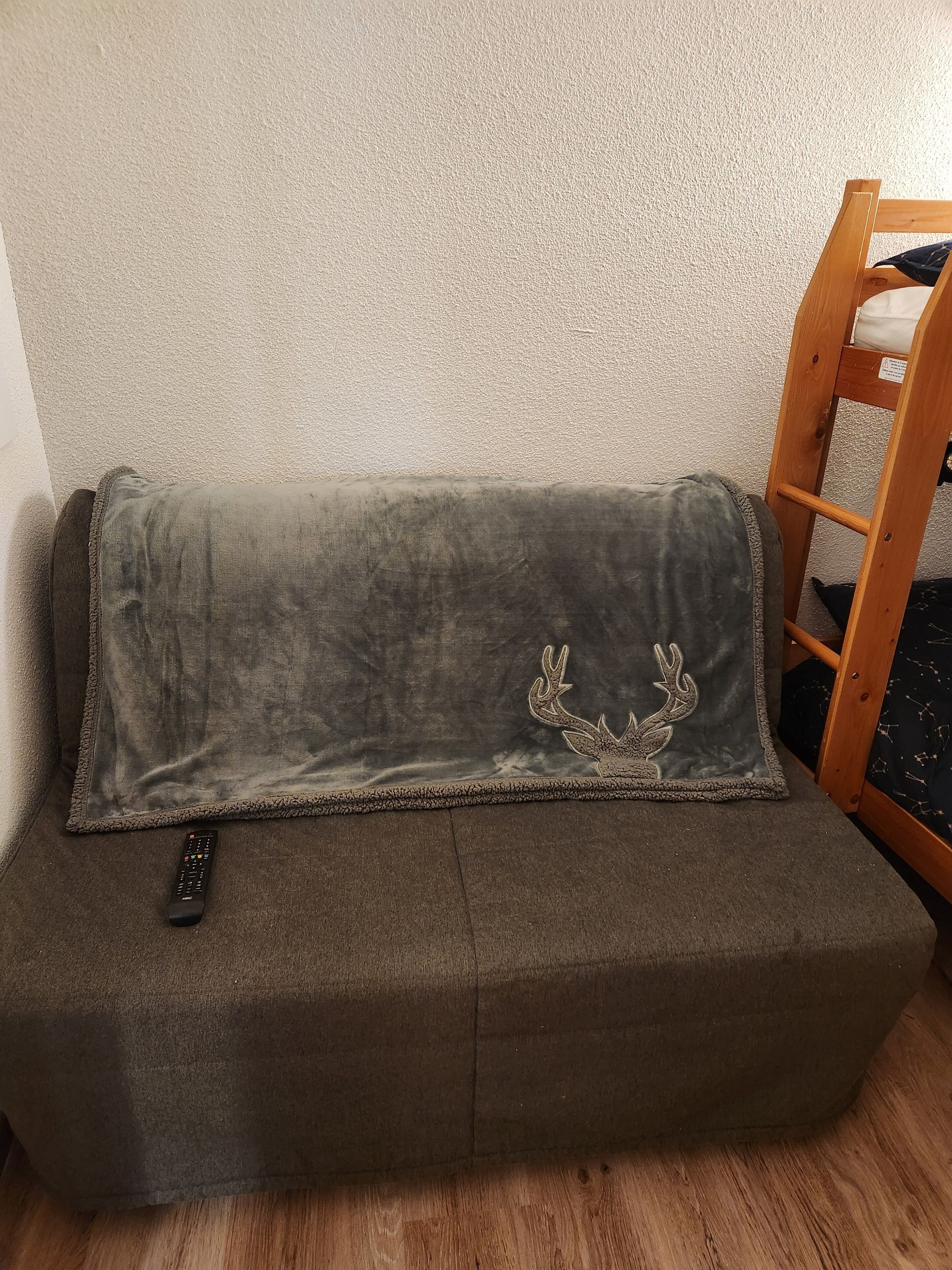 A grey sofa with a remote control on it, a wooden ladder, and a wall.