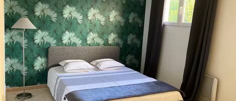A bedroom with a bed, a floor lamp, a window with curtains, and a wall with a leafy pattern.