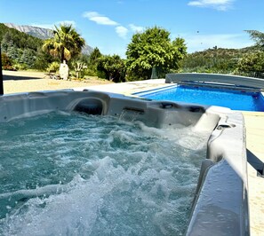 A hot tub with a pool in the background, surrounded by greenery and a clear sky.
