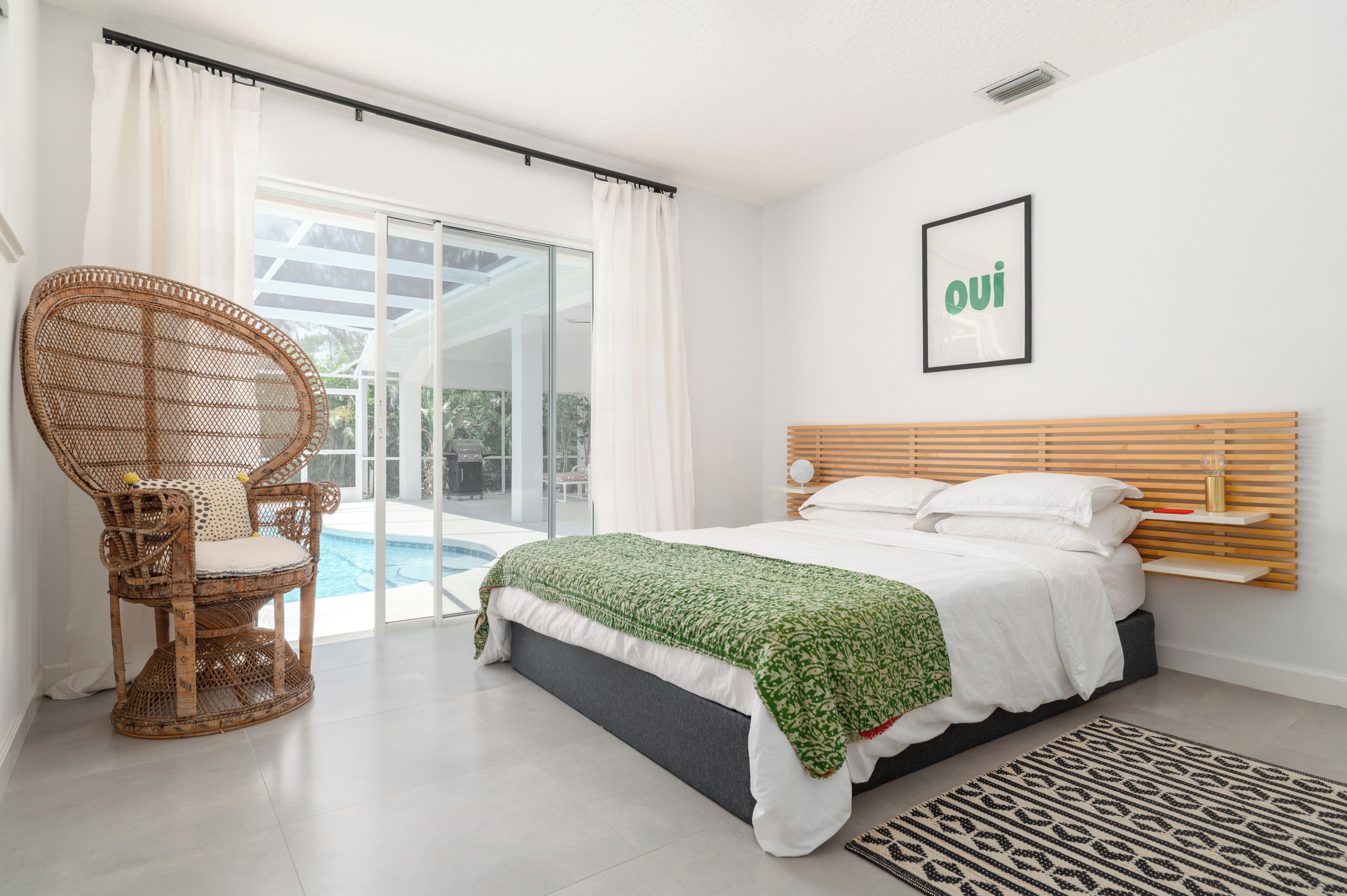 A bedroom with a bed, a wicker chair, a pool view, and a 'oui' sign on the wall.