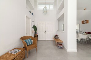 A spacious hallway with a high ceiling, a white door, wicker furniture, and a dining area with a table set for two.