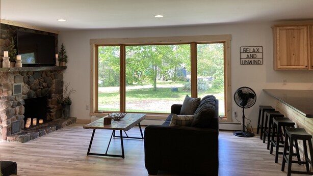 A spacious living room with a stone fireplace, a large window with a view of a yard, a flat-screen TV mounted above the fireplace, and a sign that reads "RELAX AND UNWIND".