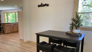 A modern kitchen with wooden cabinets, a dark wooden table, and two black barstools. A 'welcome' sign is on the wall, and a small potted plant is on the table.
