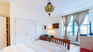 Un dormitorio con cama, escritorio, silla, lámpara, ventana con cortinas e iluminación en el techo.